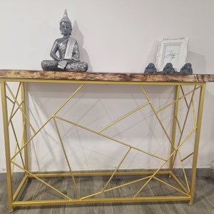 Puede incluir: Una mesa consola de metal dorado con un dise&ntilde;o geom&eacute;trico. La mesa tiene una tapa de madera y est&aacute; decorada con una estatua de Buda de plata y tres peque&ntilde;as estatuas grises.
