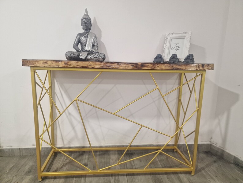 Puede incluir: Una mesa consola de metal dorado con un dise&ntilde;o geom&eacute;trico. La mesa tiene una tapa de madera y est&aacute; decorada con una estatua de Buda de plata y tres peque&ntilde;as estatuas grises.