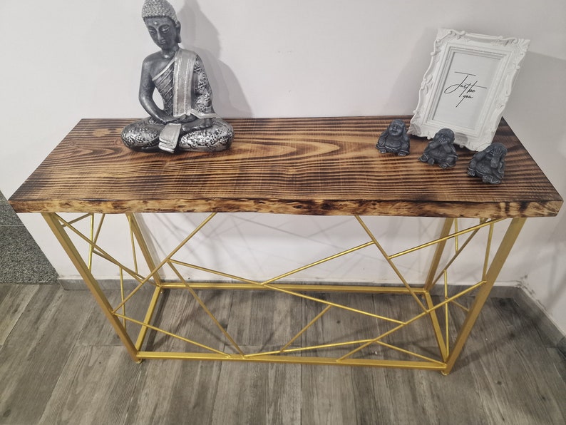 Puede incluir: Una mesa consola de madera con un marco de metal dorado. La mesa tiene una tapa de madera envejecida y un dise&ntilde;o geom&eacute;trico en el marco. Hay tres peque&ntilde;as estatuas de Buda en la mesa y un marco de imagen blanco con el texto "Just be you" en &eacute;l.