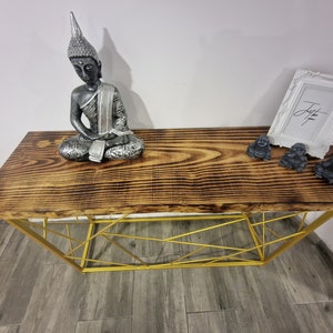 Puede incluir: Una mesa consola de madera con un marco de metal dorado. La superficie de la mesa tiene un acabado r&uacute;stico de madera quemada. Hay tres peque&ntilde;as estatuas de Buda en la mesa y un marco de imagen blanco con la palabra "Just" escrita en &eacute;l.