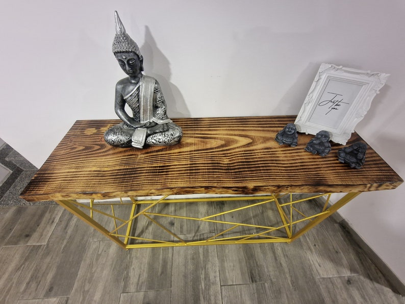 Puede incluir: Una mesa consola de madera con un marco de metal dorado. La superficie de la mesa tiene un acabado r&uacute;stico de madera quemada. Hay tres peque&ntilde;as estatuas de Buda en la mesa y un marco de imagen blanco con la palabra "Just" escrita en &eacute;l.
