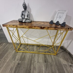 Puede incluir: Una mesa consola de metal dorado con una tapa de madera. La mesa tiene un dise&ntilde;o geom&eacute;trico y est&aacute; decorada con una estatua de Buda de plata y tres peque&ntilde;as figuras de mono grises.