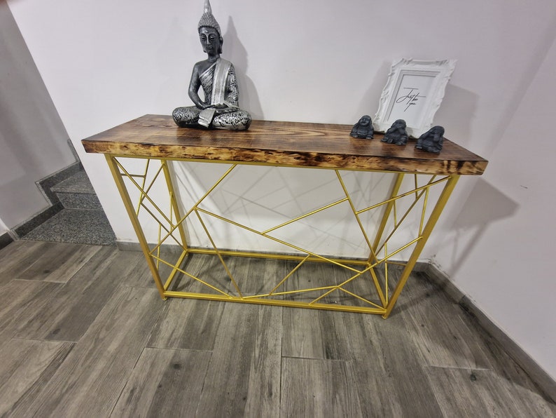 Puede incluir: Una mesa consola de metal dorado con una tapa de madera. La mesa tiene un dise&ntilde;o geom&eacute;trico y est&aacute; decorada con una estatua de Buda de plata y tres peque&ntilde;as figuras de mono grises.