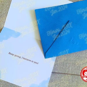 Puede incluir: Un sobre azul con el texto "Koruri_XI Jan 2024" repetido en el frente. El sobre est&aacute; abierto y el interior de la tarjeta es visible. La tarjeta tiene un fondo de cielo azul con nubes blancas y el texto "Keep going, I believe in you!" en letras marrones.
