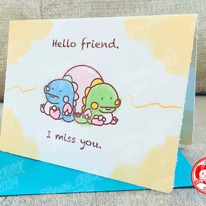 Peut inclure: Une carte de vœux illustrant un dinosaure bleu clair et vert. La carte a un fond aquarelle jaune et orange et le texte "Hello friend. I miss you."