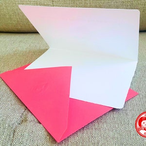 Puede incluir: Una tarjeta blanca en blanco con un sobre rosa. El sobre tiene un dise&ntilde;o en relieve en la parte delantera. La tarjeta est&aacute; doblada por la mitad y el sobre est&aacute; doblado sobre la tarjeta. La tarjeta y el sobre est&aacute;n sobre una superficie de tela beige. La imagen tambi&eacute;n incluye un sello rojo con una imagen de dibujos animados de dos animales y el texto "愛你" en chino.