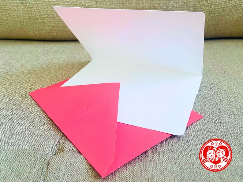 Puede incluir: Una tarjeta blanca en blanco con un sobre rosa. El sobre tiene un dise&ntilde;o en relieve en la parte delantera. La tarjeta est&aacute; doblada por la mitad y el sobre est&aacute; doblado sobre la tarjeta. La tarjeta y el sobre est&aacute;n sobre una superficie de tela beige. La imagen tambi&eacute;n incluye un sello rojo con una imagen de dibujos animados de dos animales y el texto "愛你" en chino.