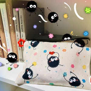 Può includere: Un astuccio con cerniera color crema con un motivo ripetuto di sprite di fuliggine neri animati con grandi occhi, che tengono stelle e cuori. Accenti di stelle e fiori colorati circondano l'astuccio. I libri sono visibili sullo sfondo.