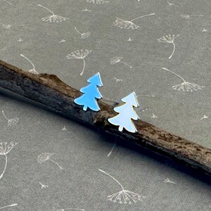 May include: A pair of tree-shaped earrings, one light blue and one white, are presented on a brown twig. The earrings have a silver outline and are set against a grey background with a subtle floral pattern. These earrings are a nature-inspired accessory.