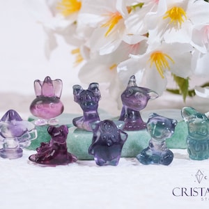 May include: A collection of eight small, colourful, crystal figurines in various animal shapes, including a unicorn, a cat, a rabbit, and a dog. The figurines are made of clear crystal with purple, blue, and pink hues. The figurines are arranged on a green surface with a white background.