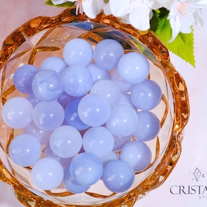 May include: A bowl filled with light blue, smooth, round, polished stones. The bowl is made of clear glass with a gold rim.