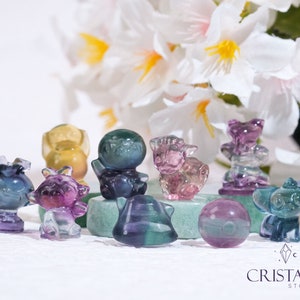 Könnte beinhalten: Eine Sammlung kleiner, bunter Kristall-Tierfiguren. Die Figuren sind aus Fluorit und in verschiedenen Farben gehalten, darunter Lila, Grün, Blau und Gelb. Die Figuren sind auf einer grünen Oberfläche mit weißem Hintergrund angeordnet.