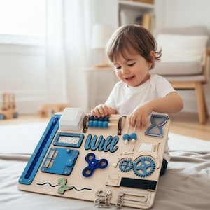 Könnte beinhalten: Ein hölzernes Beschäftigungsbrett mit verschiedenen sensorischen Aktivitäten, darunter ein blauer Fidget Spinner, blaue Zahnräder, ein Abakus mit blauen Perlen, ein Lichtschalter, ein blauer Traktor und der Name Will in blauer Schreibschrift.