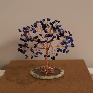 May include: A decorative tree sculpture featuring a twisted copper wire trunk and branches adorned with numerous small, polished deep blue lapis lazuli stone chips. The tree is anchored to a flat, circular, light-coloured stone base.