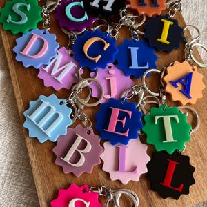 May include: A collection of colourful acrylic keychains, each featuring a scalloped, star-shaped base in various solid colours with a contrasting raised letter in the centre. Each keychain includes a silver-toned metal split ring and a lobster clasp.