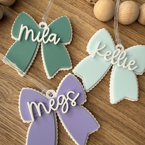 May include: Three bow-shaped name tags in shades of green, blue, and purple, each with a white scalloped edge. The names "Mila", "Kellie", and "Megs" are written in white script across the bows. Clear plastic loops are attached for hanging.