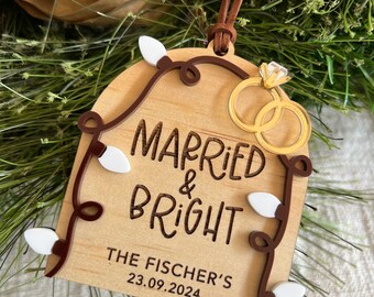 May include: Wooden wedding ornament with the words "Married & Bright" and "The Fischer's 23.09.2024". It features a string of lights and two gold wedding rings. A brown leather cord is attached for hanging.