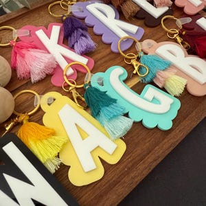 May include: A selection of personalised keyrings, each featuring a letter design. The keyrings have different coloured bases, white letters, and matching tassels. The keyrings are displayed on a wooden tray, showcasing a variety of colours and styles.
