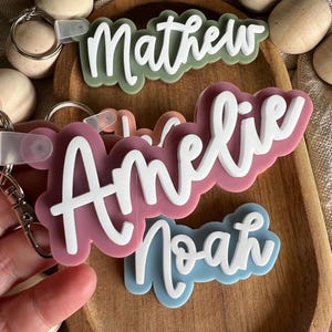 Puede incluir: Tres llaveros personalizados con los nombres "Mathew", "Amelie" y "Noah" escritos en blanco sobre fondos acrílicos de colores. Los llaveros se muestran en una bandeja de madera con bolas de madera.