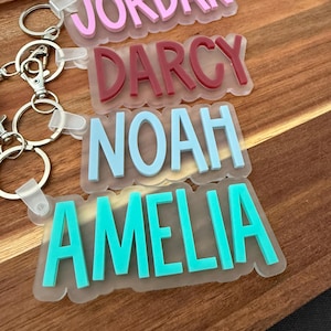 Könnte beinhalten: Vier personalisierte Schlüsselanhänger mit erhabenen, farbigen Acrylbuchstaben auf einer mattierten Acrylbasis. Die Namen Jordan in Pink, Darcy in Kastanienbraun, Noah in Hellblau und Amelia in Türkis sind an silbernen Metallringen befestigt.