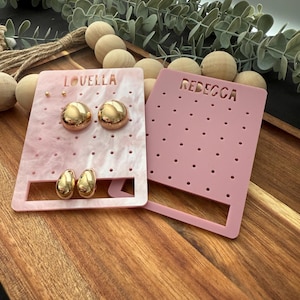 May include: Two rectangular earring display stands with perforated holes. One stand is marbled light pink with the name LOVELLA and holds two pairs of gold earrings. The other stand is solid matte pink with the name REBECCA and is empty.