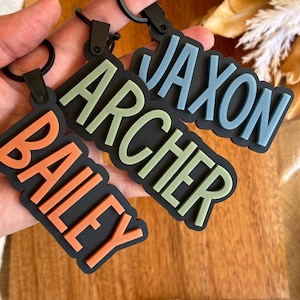 Peut inclure: Trois porte-clés personnalisés avec des lettres en relief orange, vert sauge et bleu poussiéreux sur une base noire. Chaque porte-clés affiche un nom : Bailey, Archer ou Jaxon. Ils sont fixés à des anneaux et des clips en métal noir.