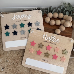 May include: Two wooden reward charts with names, star decorations, and reward sections. One features navy, teal, and silver glitter stars, the other has pink and gold glitter stars. Both have white text and a white reward area.