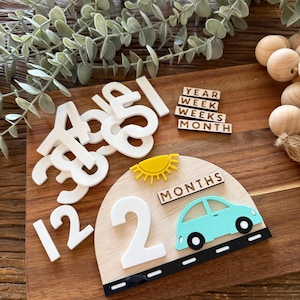 May include: A wooden milestone plaque shaped like a semi-circle featuring a yellow sun, a light blue car, and the text MONTHS. It includes a set of white acrylic numbers and small wooden tiles with the words YEAR, WEEK, WEEKS, and MONTH.