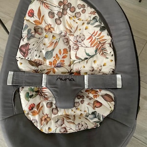 Pode incluir: Um balanço para bebé cinzento com um almofada estampada floral branca e castanha. O balanço tem um exterior de tecido cinzento e uma correia branca com a palavra "Nuna" impressa nela.