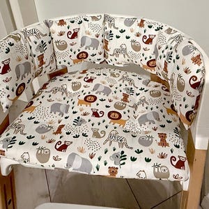 Può includere: Una sedia alta bianca con un sedile imbottito e cuscini per lo schienale. I cuscini sono rivestiti in un tessuto bianco con un motivo colorato di animali, tra cui elefanti, bradipi e leoni.