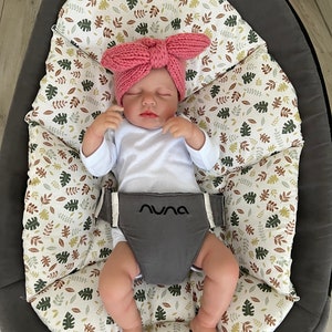 Könnte beinhalten: Ein Baby mit einem rosa Schleifenhaarband und einem weißen Strampler sitzt in einem grau-weißen gemusterten Baby-Schaukelstuhl. Der Schaukelstuhl hat den Schriftzug "Nuna" aufgedruckt.