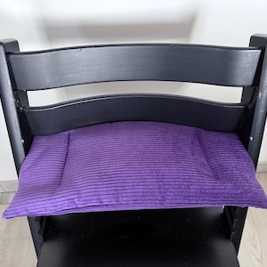 Op de afbeelding: Een zwarte houten stoel met een gebogen rugleuning en een rechthoekig, paars corduroy kussen. Het kussen is gecentreerd op de zitting en biedt een contrasterende kleur tegen het donkere hout.