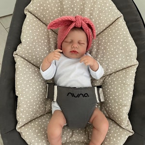 May include: A baby wearing a pink knitted headband with a bow, lying on a beige and white polka dot patterned cushion in a gray baby bouncer. The bouncer has the word "NUNA" printed on the front.