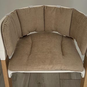 May include: A white high chair with a beige corduroy cushion and back support. The cushion has a quilted design and the back support has four separate cushions.