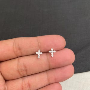 Könnte beinhalten: Ein Paar kleine, silberfarbene Kreuz-Ohrstecker. Jeder Ohrring hat ein Kreuzdesign mit kleinen, klaren Edelsteinen. Die Ohrringe werden auf einer hellen Oberfläche präsentiert.