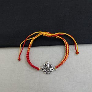 May include: A braided cord bracelet featuring a silver-toned metal charm of a Ganesha figure. The bracelet is constructed from intertwined red and yellow threads and includes an adjustable sliding knot closure with loose cord ends.