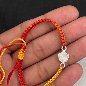 May include: A handmade bracelet featuring a woven cord in red, orange, and yellow. The bracelet has a silver-colored rose charm. The cord is knotted in a macrame style. The bracelet is displayed against a dark background.