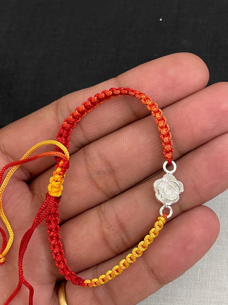 May include: A handmade bracelet featuring a woven cord in red, orange, and yellow. The bracelet has a silver-colored rose charm. The cord is knotted in a macrame style. The bracelet is displayed against a dark background.