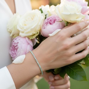 May include: A delicate gold bracelet with small, round, turquoise-colored stones encircles a wrist. The person is holding a bouquet of white roses and pink peonies. The background is blurred, suggesting an outdoor setting.