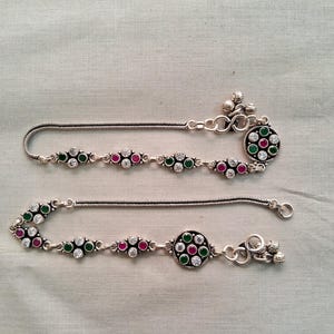 Op de afbeelding: Een zilveren enkelband met een ronde bedel met groene en roze stenen. De enkelband heeft een kettingontwerp en is versierd met kleine witte stenen.