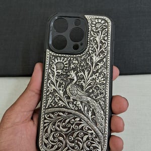 May include: A silver phone case with an intricate peacock design. The case is made of metal and has a black background. The peacock is surrounded by floral patterns.