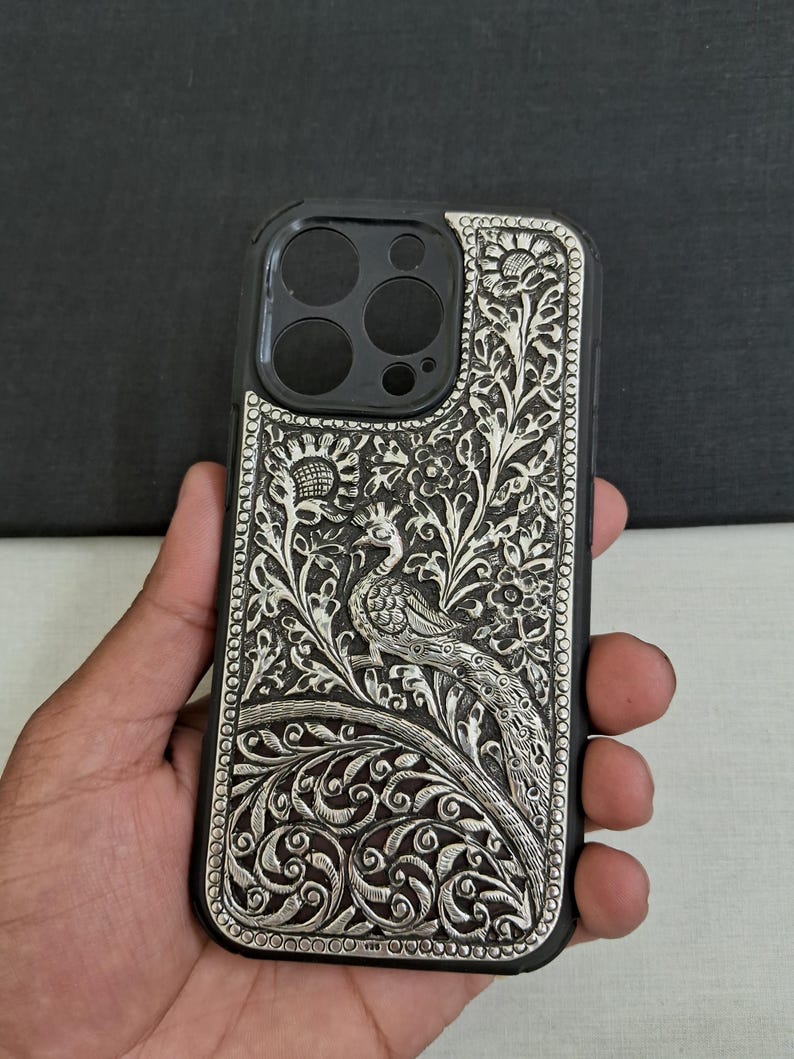 May include: A silver phone case with an intricate peacock design. The case is made of metal and has a black background. The peacock is surrounded by floral patterns.