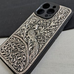 May include: A black phone case with a silver peacock design. The case has a raised edge around the camera lens.