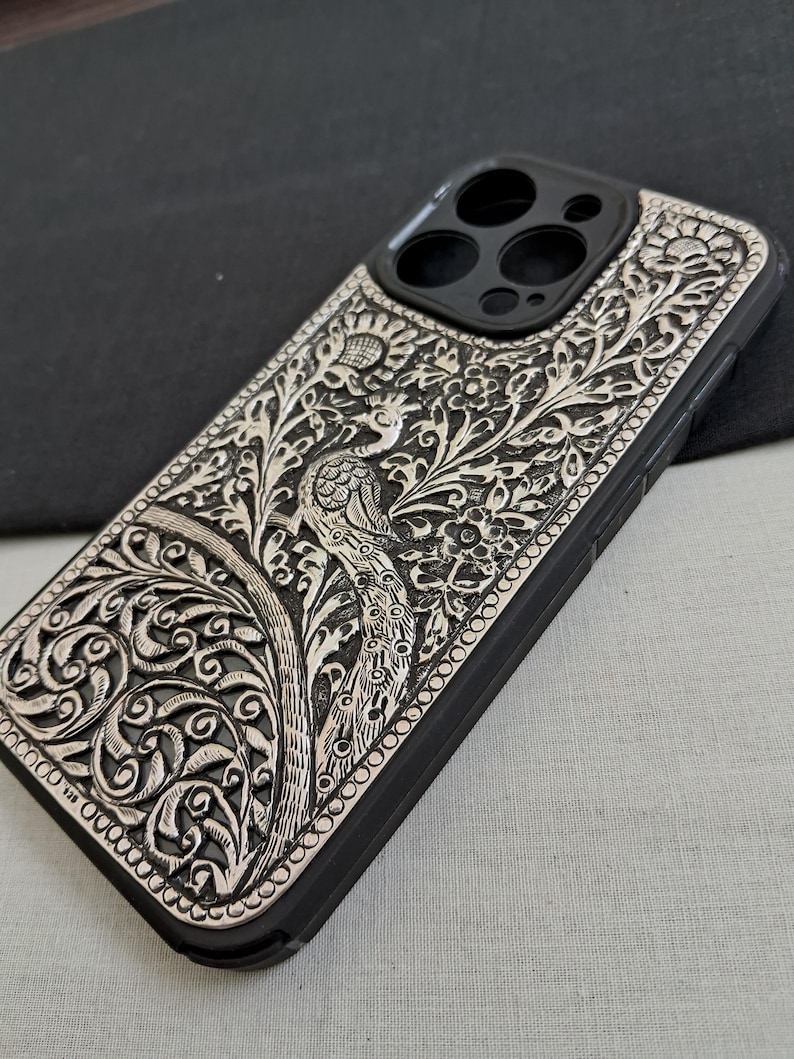 May include: A black phone case with a silver peacock design. The case has a raised edge around the camera lens.