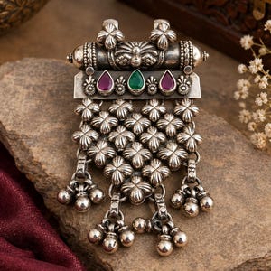 May include: An oxidized silver pendant featuring a central horizontal bar with two magenta and one green teardrop-shaped stones. The lower section consists of a triangular arrangement of small floral motifs with dangling spherical silver beads.
