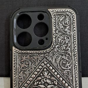 Puede incluir: Una funda de tel&eacute;fono negra con un dise&ntilde;o floral plateado. La funda tiene un recorte para el objetivo de la c&aacute;mara.