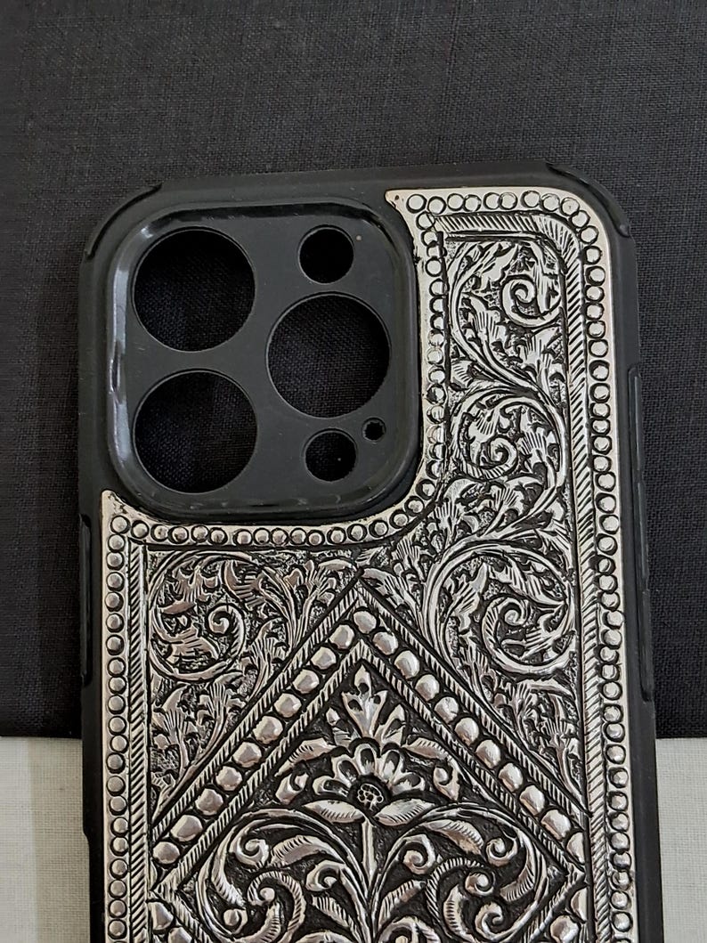 Puede incluir: Una funda de tel&eacute;fono negra con un dise&ntilde;o floral plateado. La funda tiene un recorte para el objetivo de la c&aacute;mara.