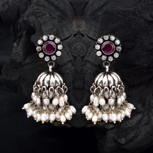 May include: A pair of silver earrings with a floral design. Each earring has a central red gemstone surrounded by small clear stones. The lower section is bell-shaped, adorned with dangling white pearls. The earrings are set against a dark background.