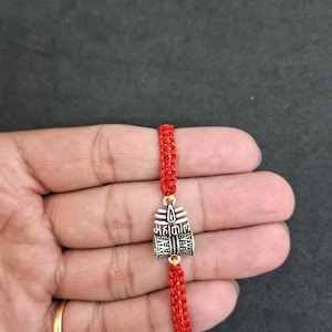 May include: Red woven bracelet with a silver-colored charm. The charm has intricate detailing and text. The bracelet is held against a dark background. The bracelet is a traditional style.