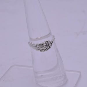 May include: A silver ring with a wing design. The ring is displayed on a clear ring stand.